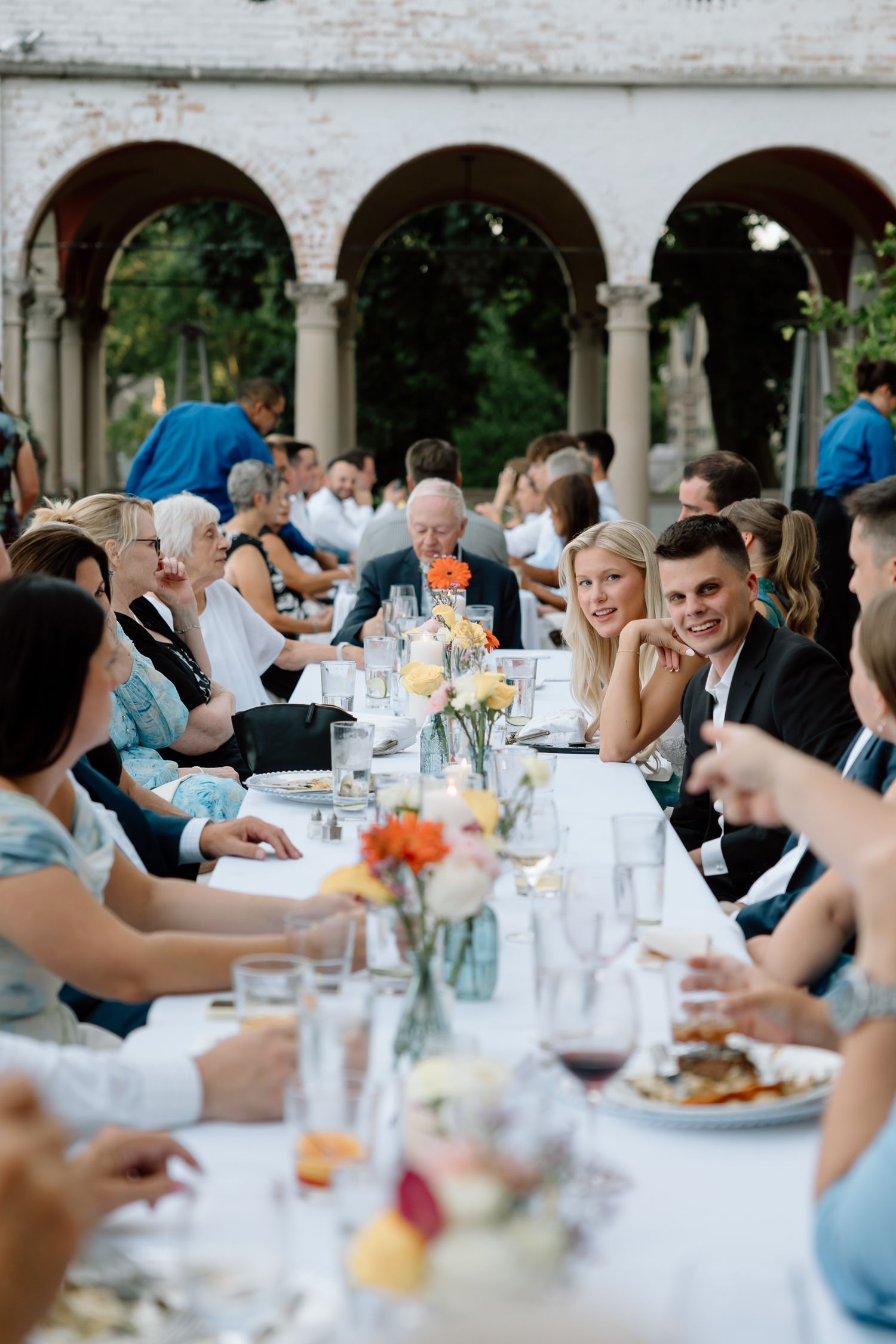 Bride and groom laughing with guests at elegant outdoor wedding reception under arched colonnade.
