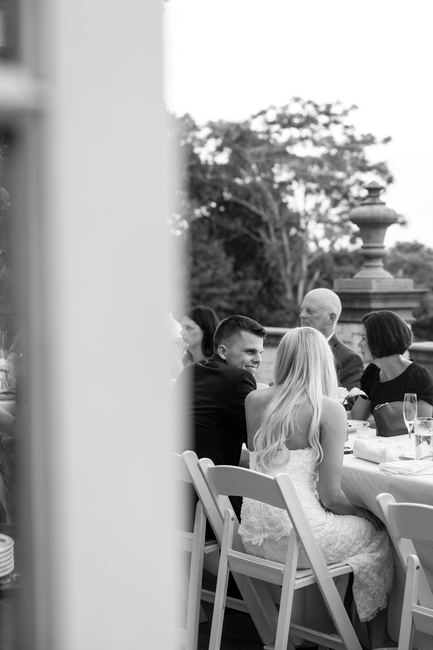 Black and white photo of bride and groom seated at outdoor wedding reception table with guests.