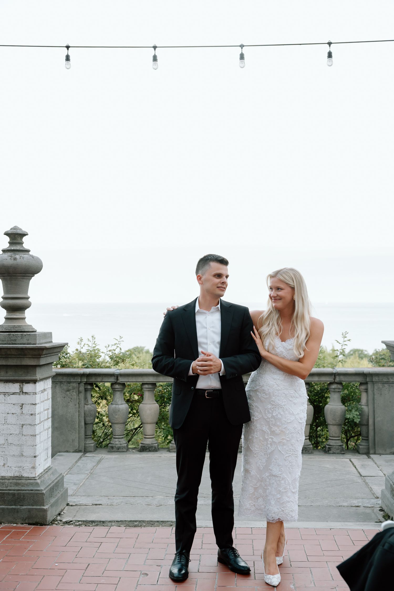 Bride and groom standing together on terrace with ocean view and string lights at sunset wedding.