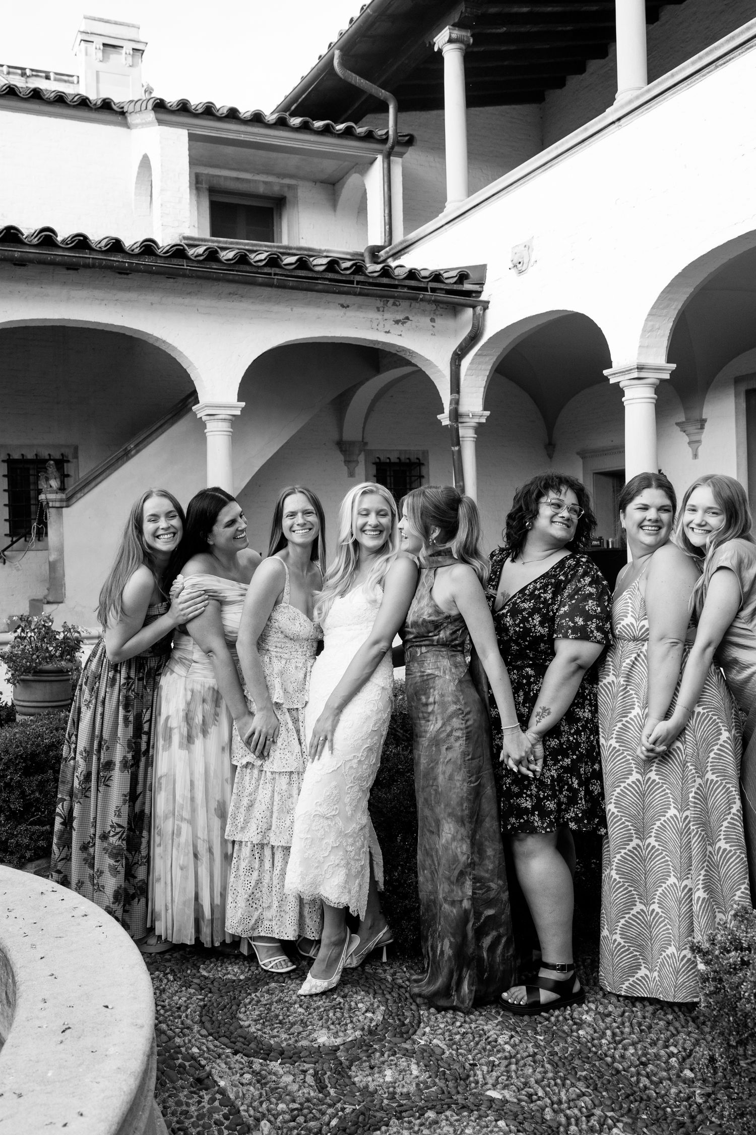 Black and white photo of a bride in white dress posing with group of women friends at elegant villa wedding venue.