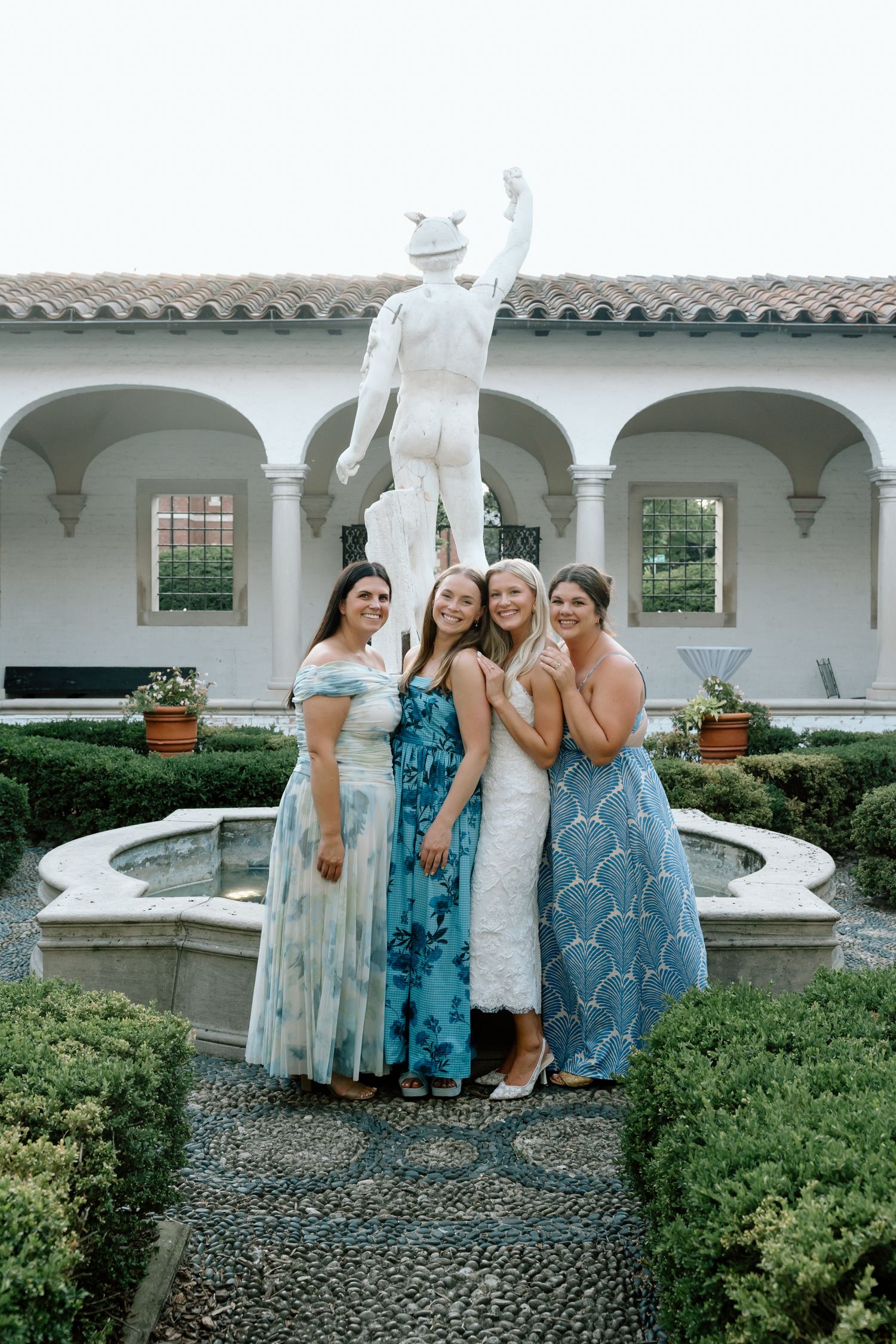 Bride posing with three bridesmaids in blue dresses by fountain statue in Italian villa garden.