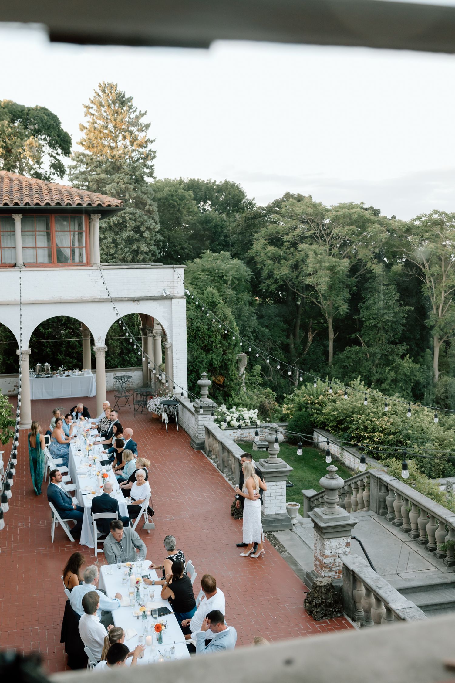 Aerial view of outdoor wedding reception on terrace with long dining tables, string lights, and lush garden backdrop.