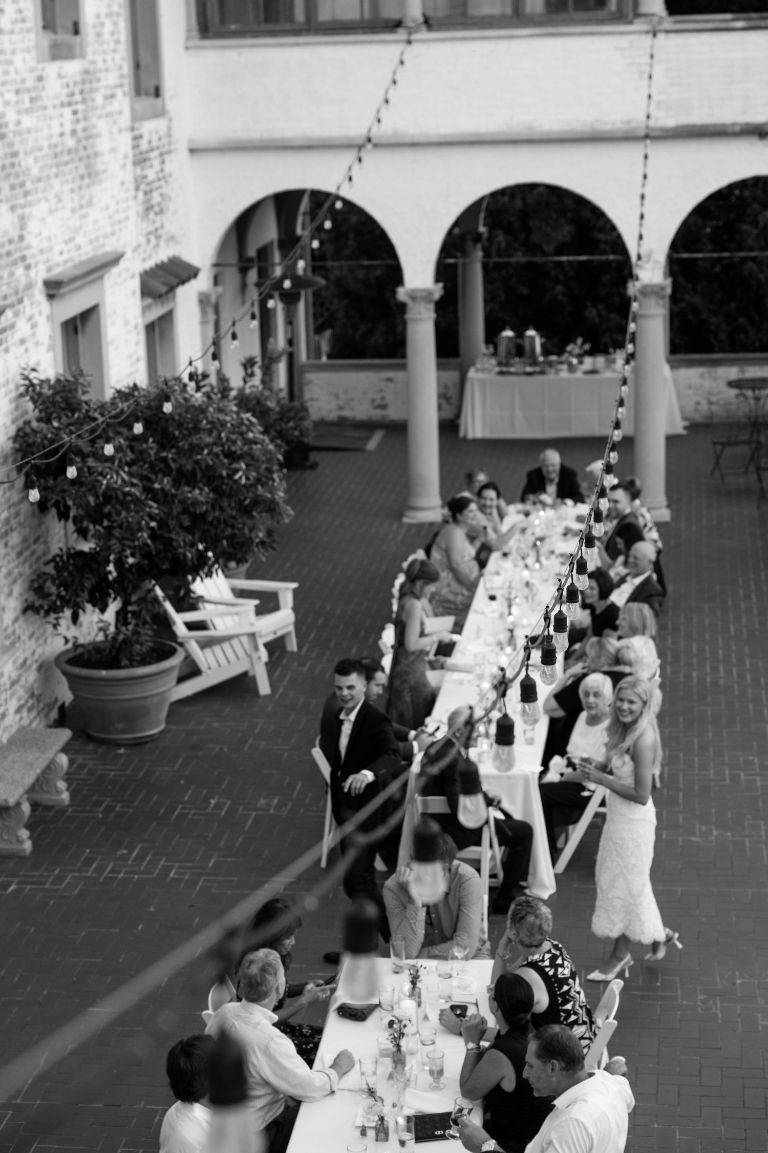 Black and white aerial view of wedding reception guests dining at long outdoor table in elegant courtyard with arched colonnade.