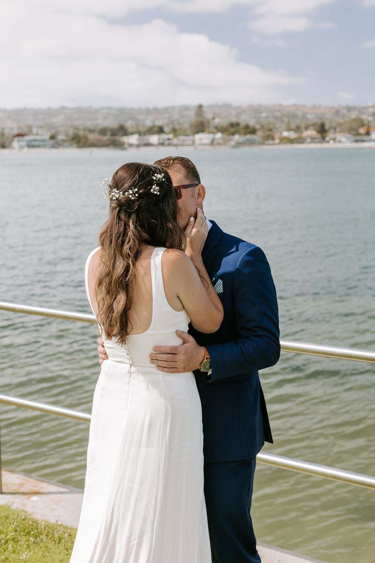 Bride and groom share a kiss by waterfront railing, bride in white gown with floral hair accessory, groom in navy suit at Tower Beach Club.