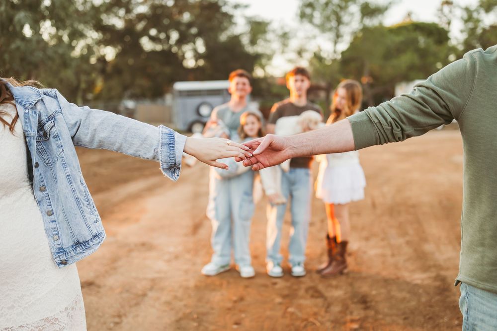 Couple holding hands on a dirt path with family members standing in the background at sunset.