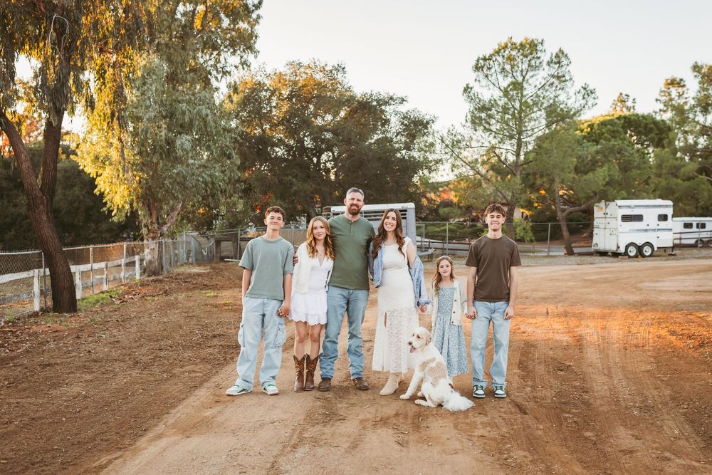 Family of six with a white dog posing on a dirt path at golden hour near a horse trailer.