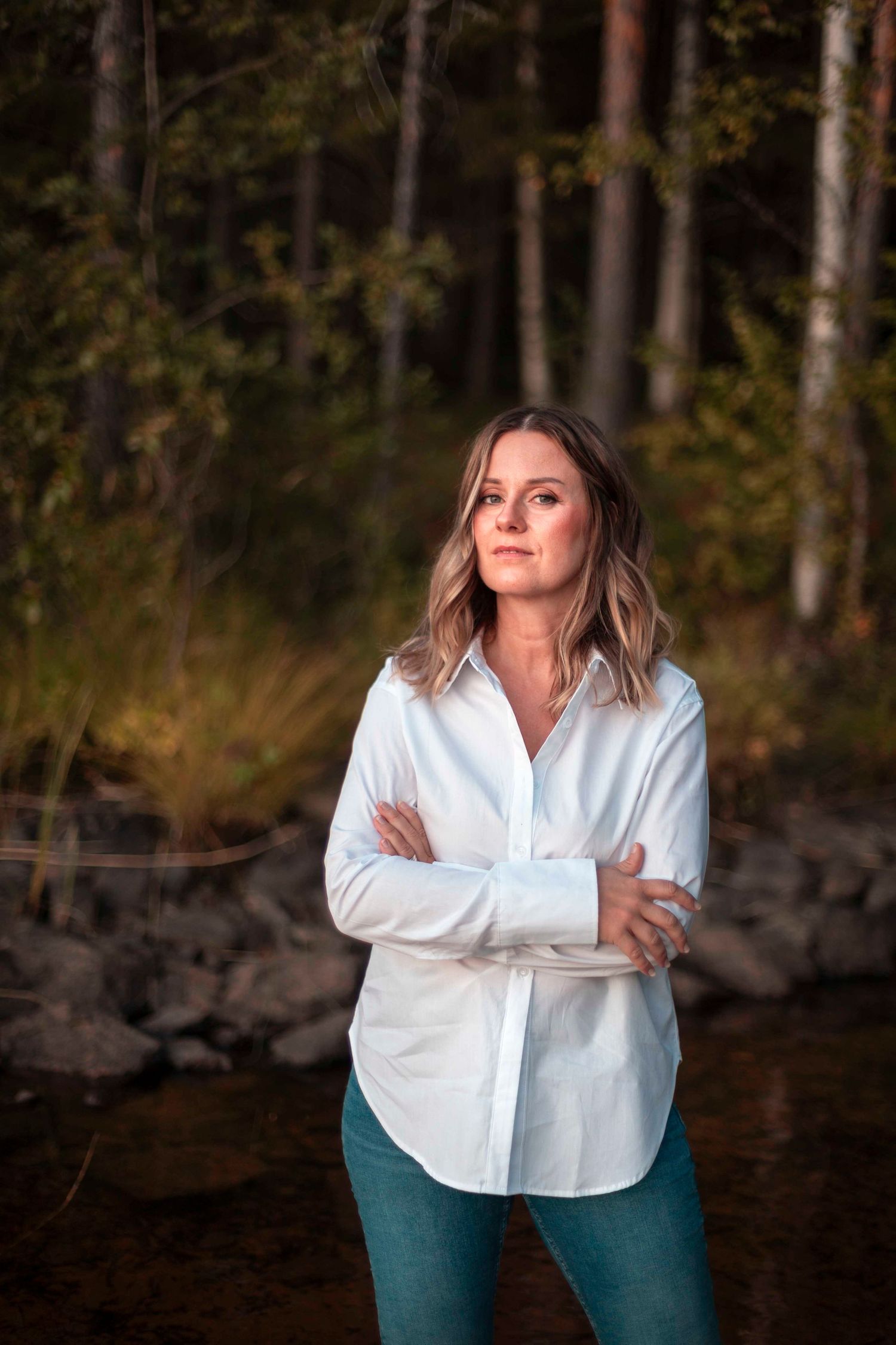 Confident woman in white shirt with arms crossed standing near a forest stream.