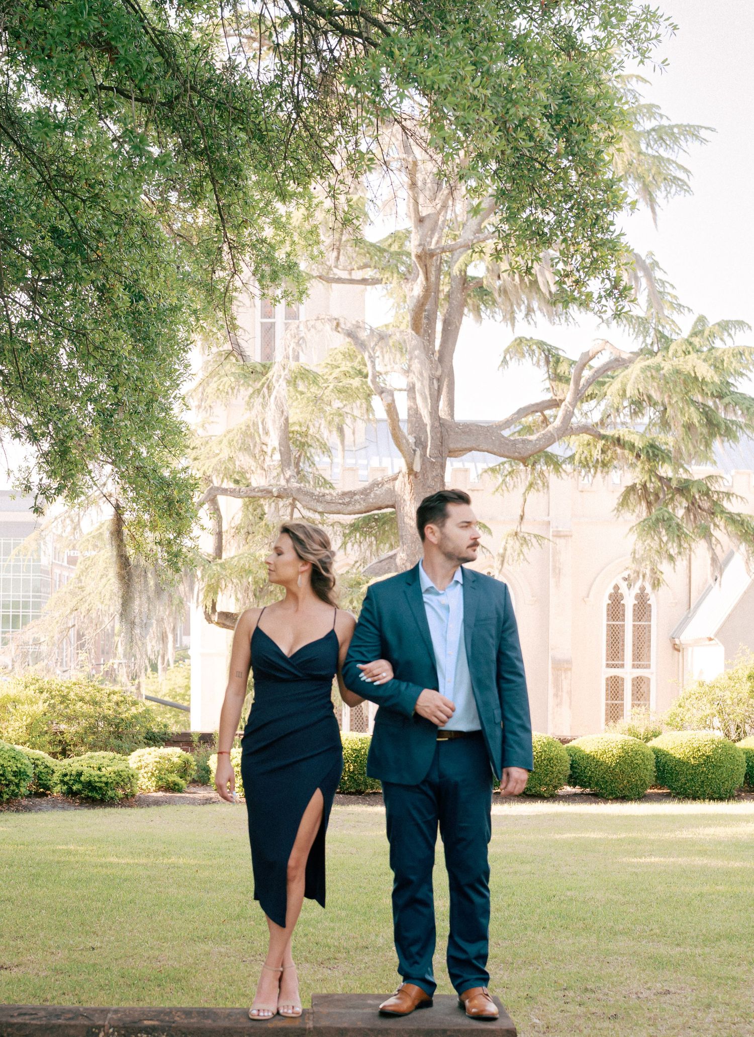Couple in elegant attire walks through lush garden near historic building, woman in black dress, man in teal suit.