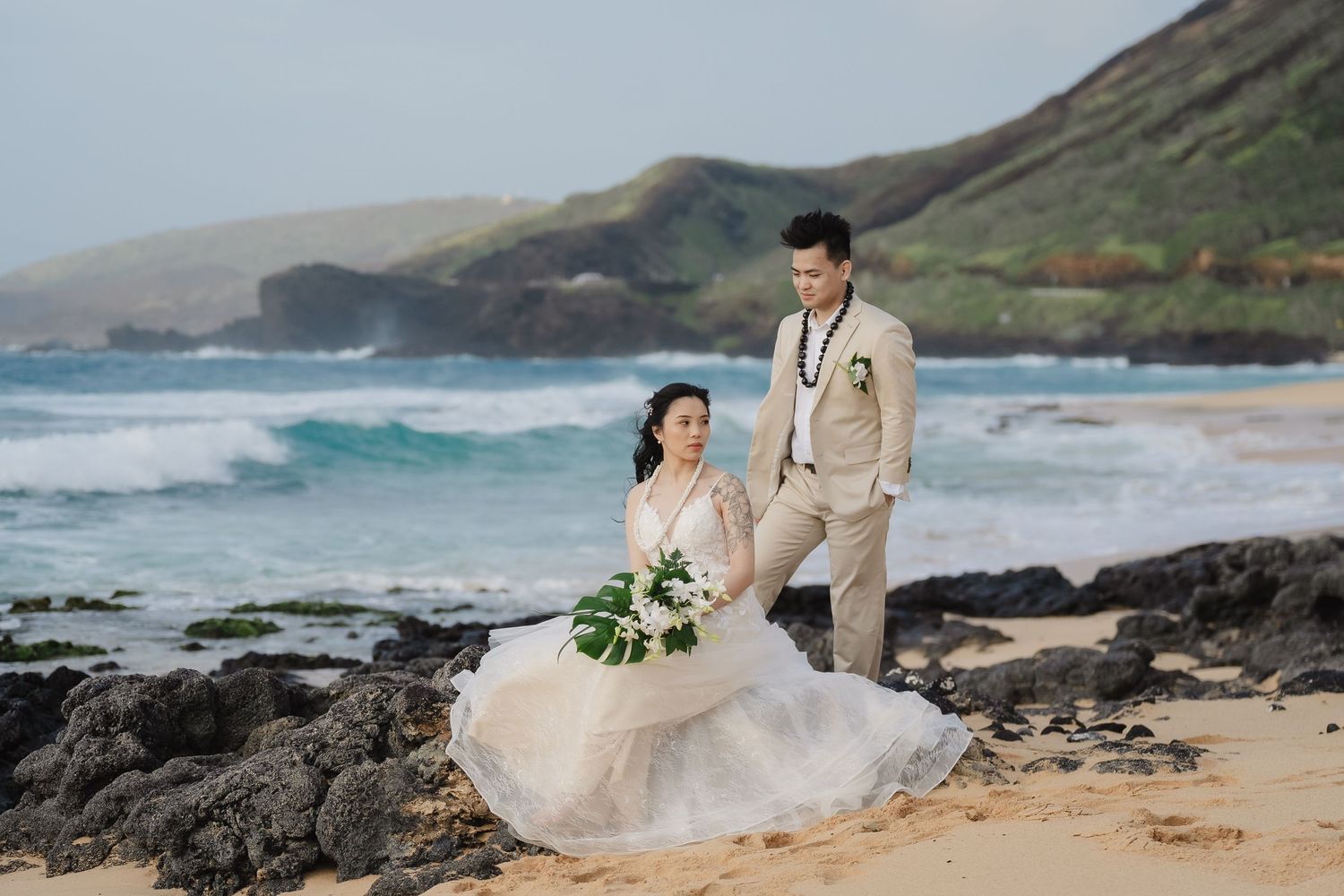 Couple in wedding attire posing on a sandy beach with rocky coastline and ocean waves in the background.