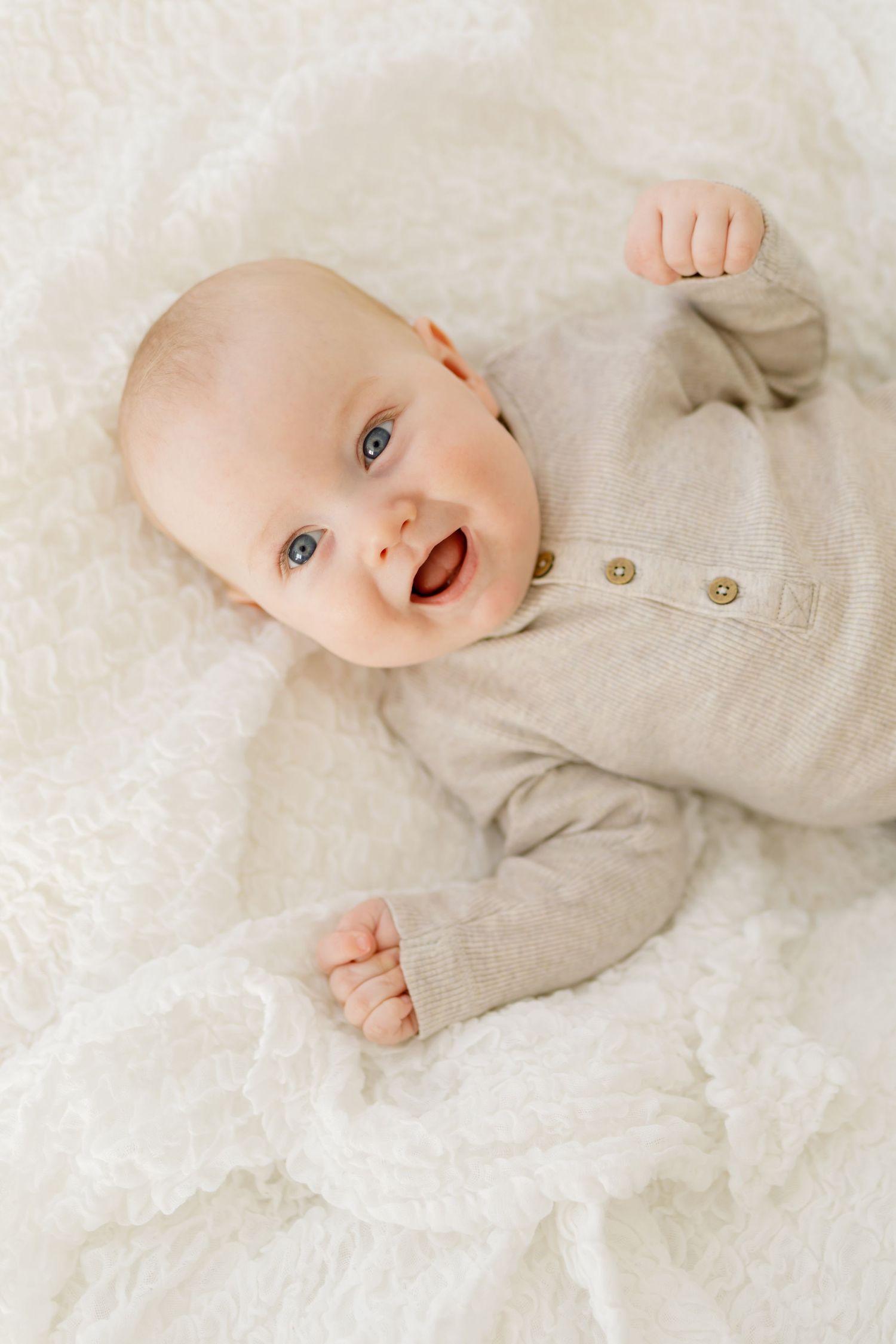 Leende baby med blå ögon klädd i en beige stickad byxa, liggande på en vit fluffig filt.