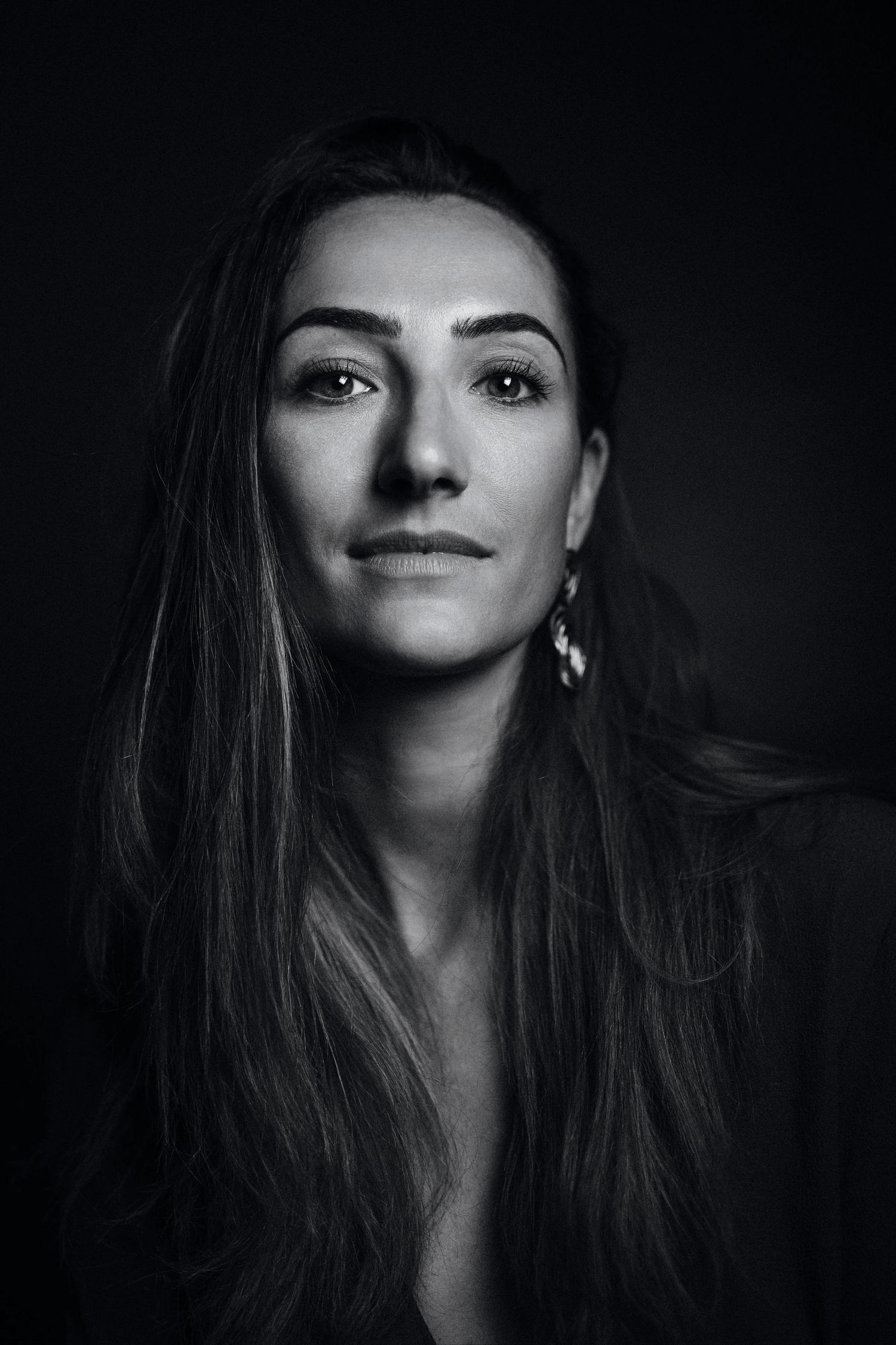 Black and white portrait of a woman with long hair and earrings against a dark background.