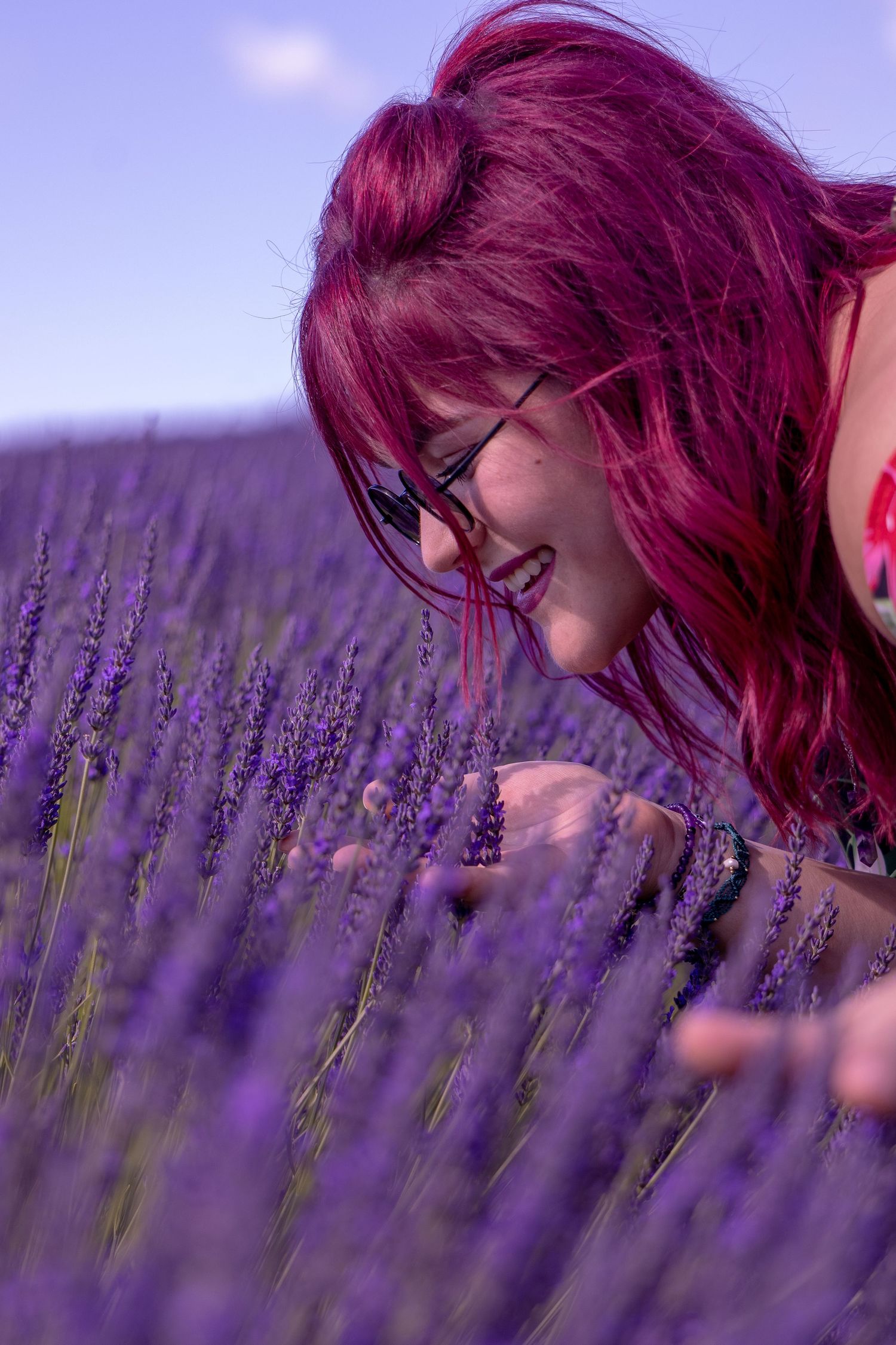 A smiling woman with red hair and glasses bends down to smell purple lavender flowers in a blooming field.