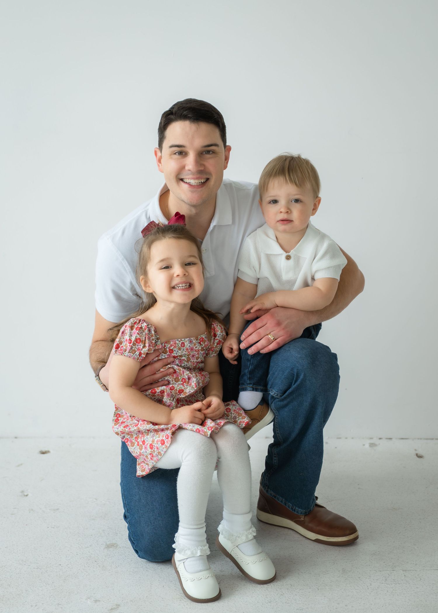 A smiling man poses with two young children in a bright studio, all dressed in casual outfits for a family portrait.