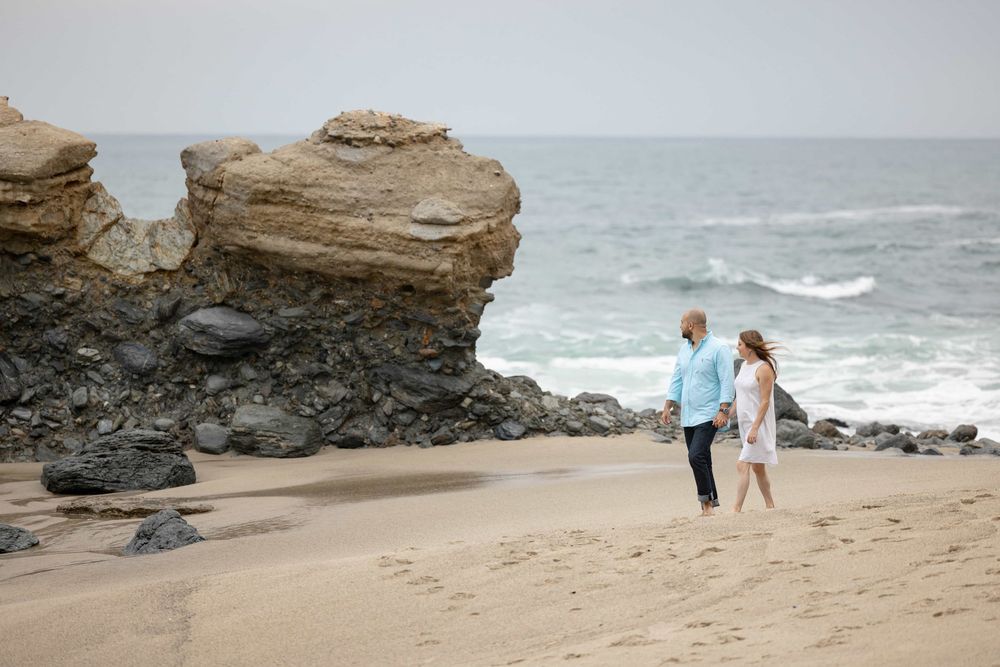 Couple walking hand in hand on a sandy beach with dramatic rock formations and ocean waves.