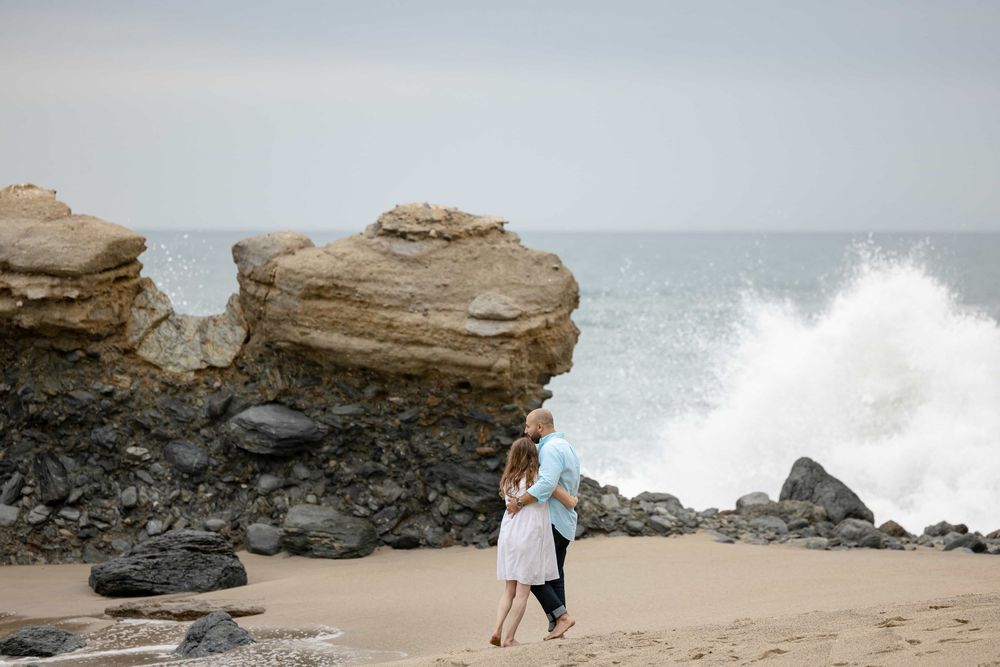 Couple embracing on beach with crashing waves and rock formations.