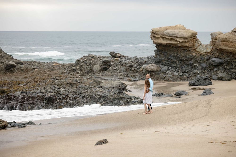 Woman in white dress standing alone on a sandy beach with rocky cliffs and ocean waves.