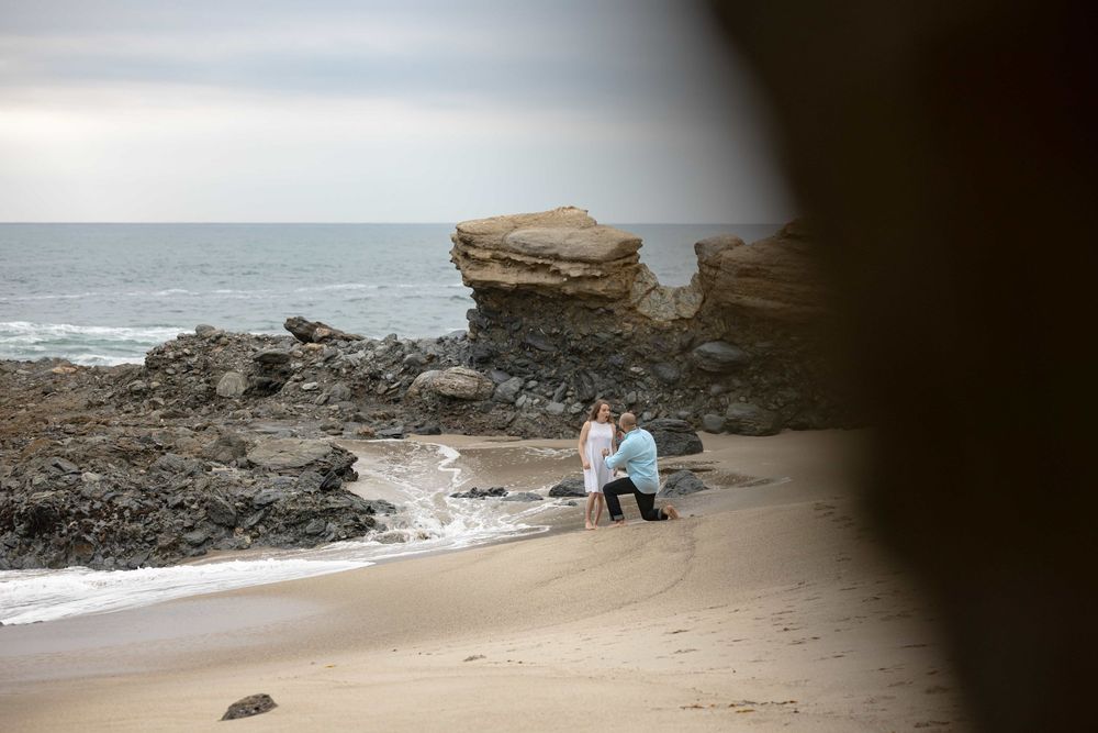 Man proposing to woman on a sandy beach with large rock formations and ocean in background.