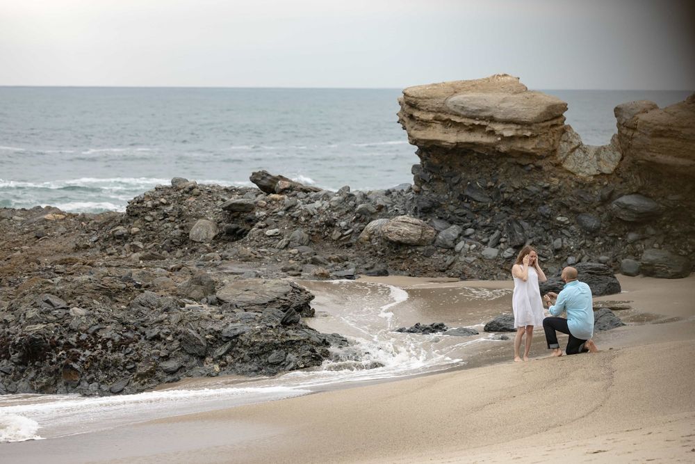 Man down on one knee proposing to woman on a beach near rocky cliffs and ocean waves.