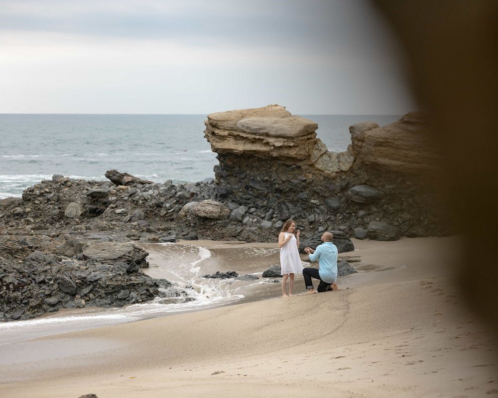 Romantic beach marriage proposal with man kneeling before woman near scenic rock formations.