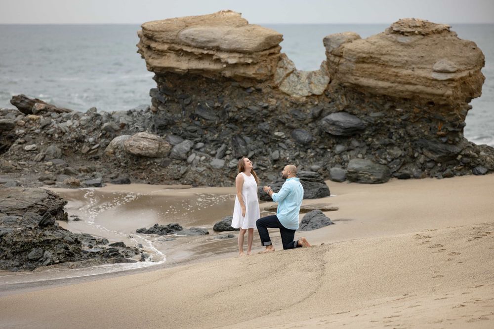 Man kneeling on one knee proposing to woman on sandy beach with large rock formations.