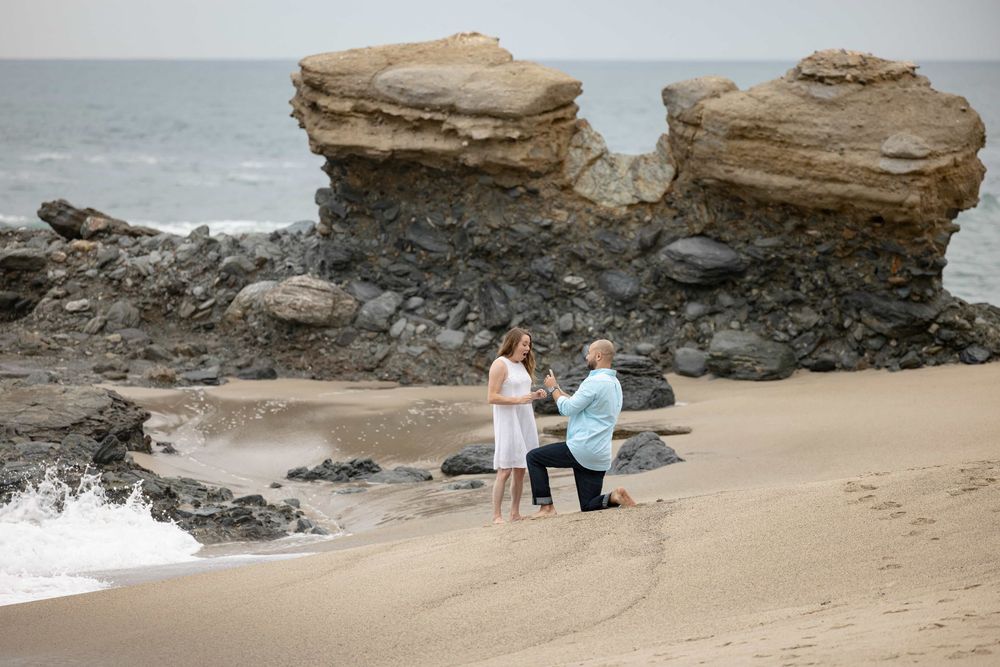 Beach marriage proposal with man on one knee before woman near dramatic coastal rock formations.
