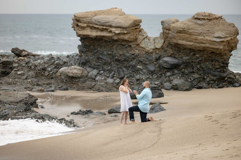 Couple sharing a romantic marriage proposal moment on sandy beach with rocky ocean backdrop.