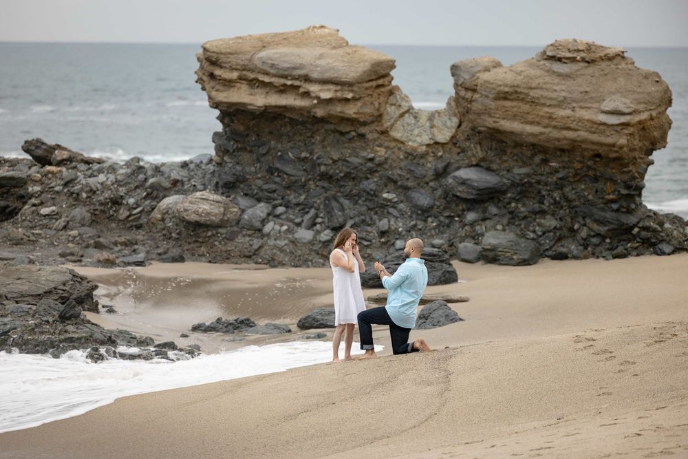 Man proposing to woman on a scenic beach with twin rock formations and ocean waves.