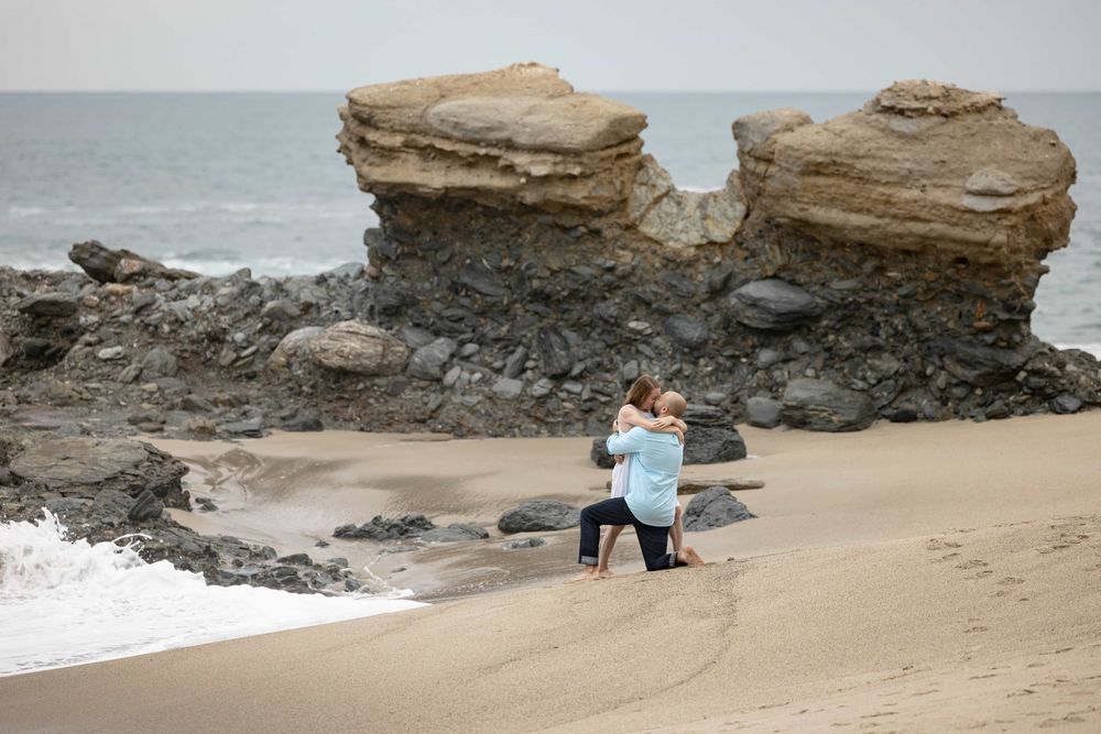 Happy couple embracing on sandy beach after marriage proposal near dramatic coastal rock formations.