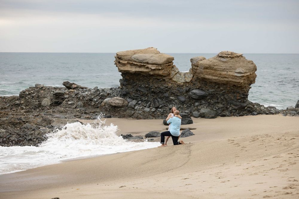 Woman kneeling on sandy beach photographing dramatic rock formations as ocean waves crash nearby.
