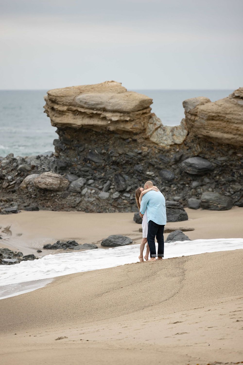 Couple embracing on a sandy beach near large rock formations with ocean waves in the background.