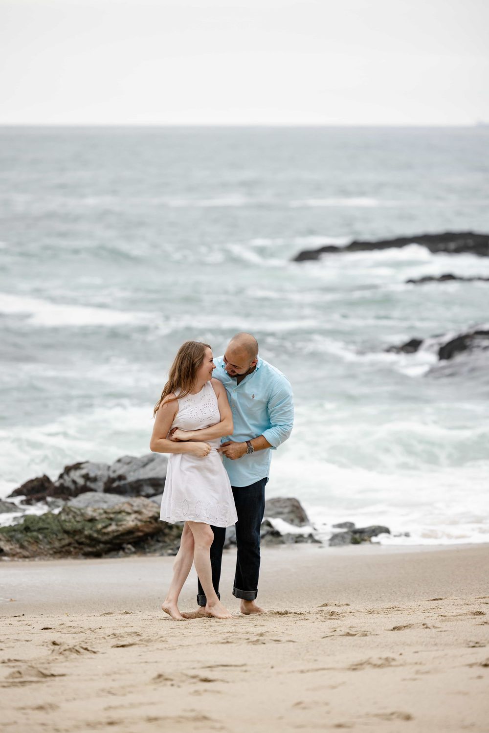 Happy couple walking barefoot on sandy beach with turquoise ocean waves and rocky shoreline behind them.