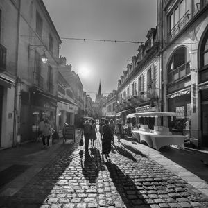 Black and white street photo of pedestrians walking on cobblestone street with sunlight and long shadows.