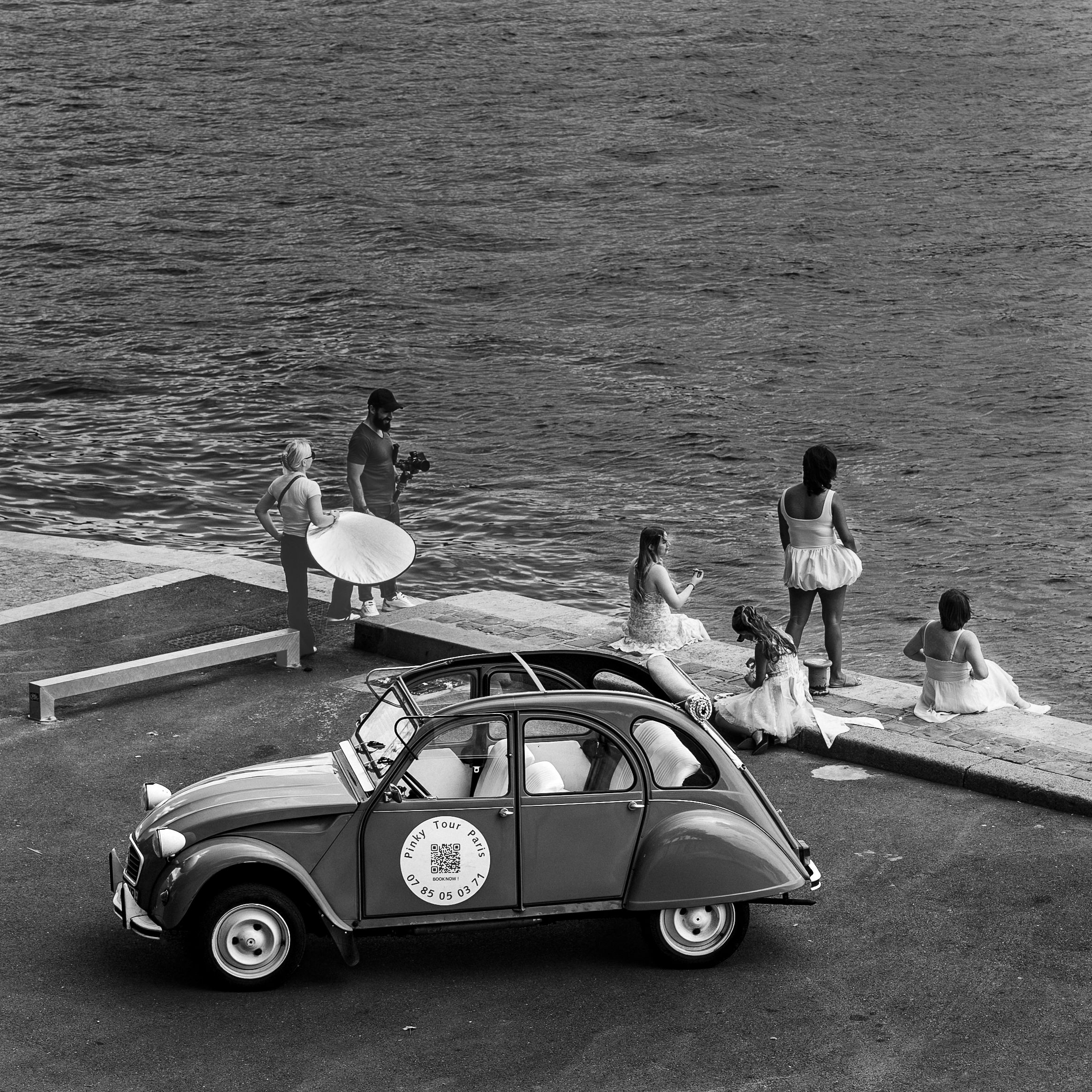 Vintage black and white photo of a Citroen 2CV parked near a waterfront with people in the background.