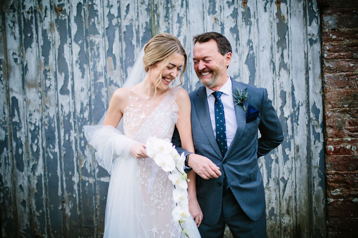 Happy bride and groom laughing together in wedding attire against a rustic weathered wall backdrop.