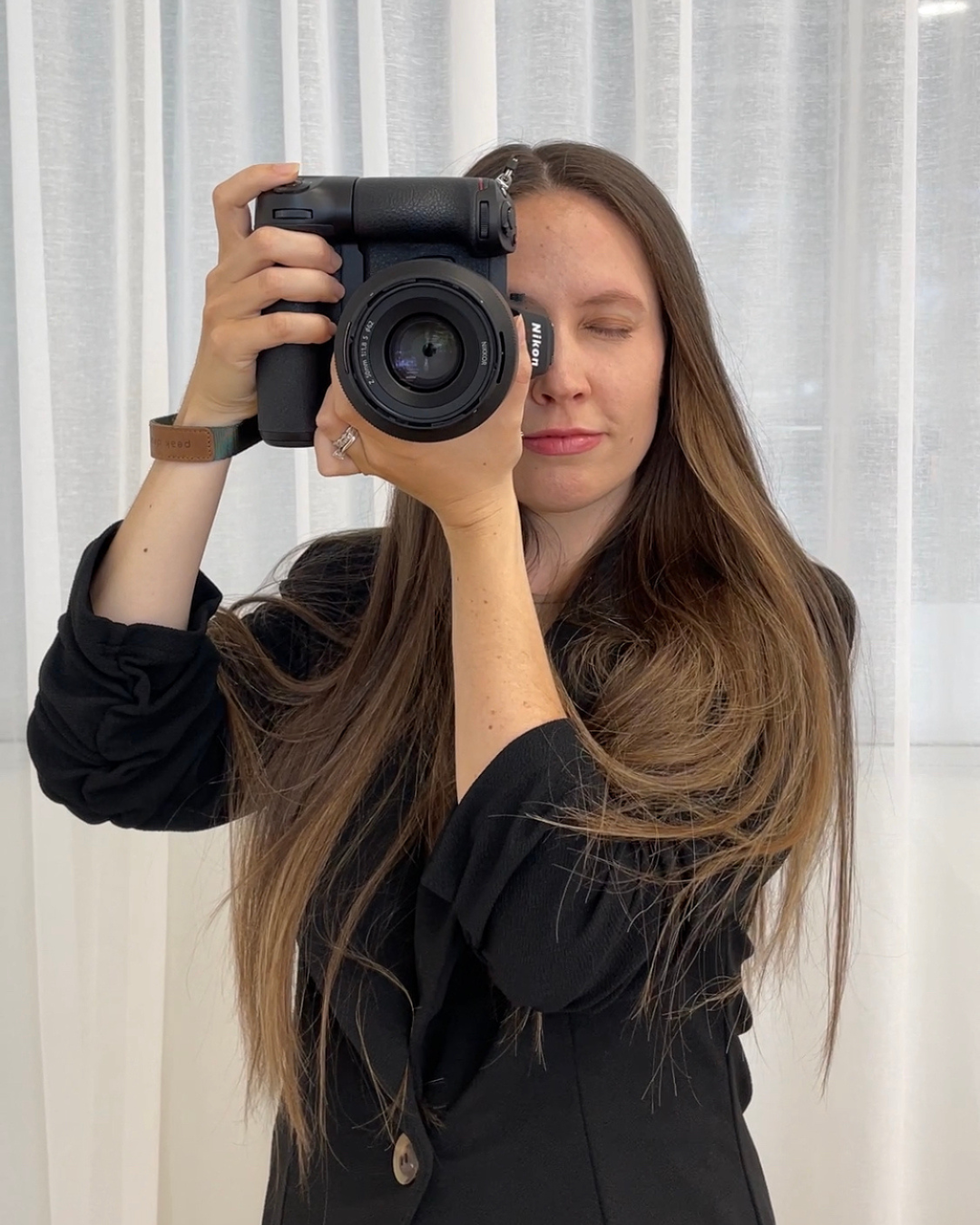 Woman with long brown hair holding a black DSLR camera up to her eye, wearing a black outfit indoors.