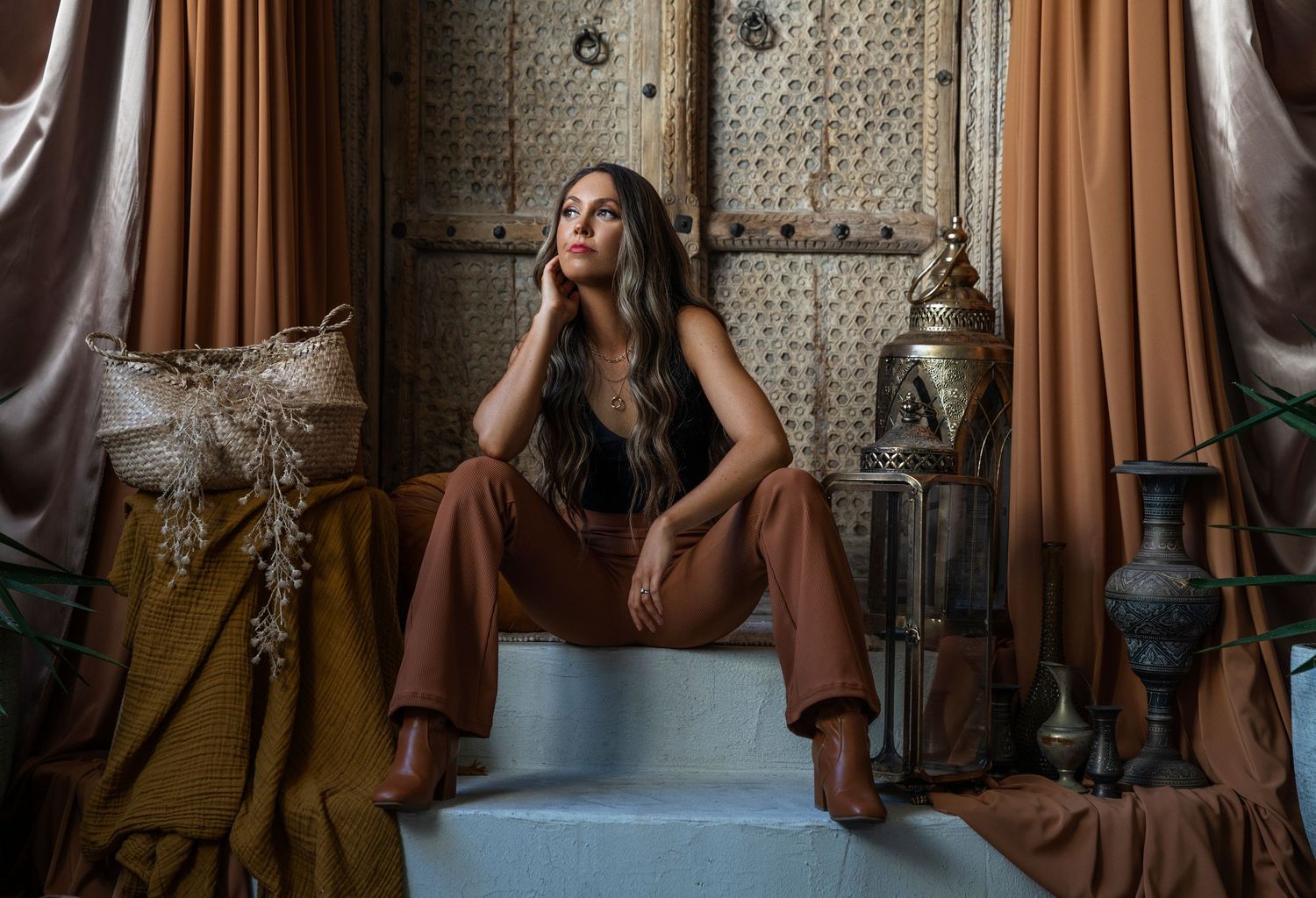 Woman in brown wide-leg pants poses elegantly in a Moroccan-inspired room with ornate decor and draped curtains.