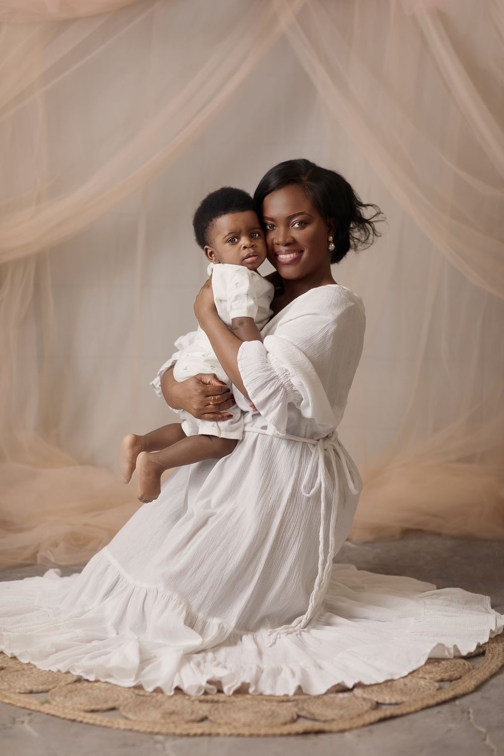 A smiling Black mother in a white dress holds her baby in a white outfit against a soft beige draped backdrop.