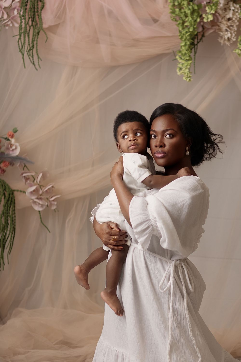 A Black mother in a white dress holds her baby in a floral studio setting.