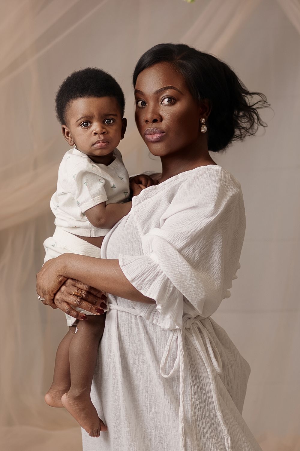 A Black mother in a white dress holds her baby boy dressed in white against a soft neutral studio background.