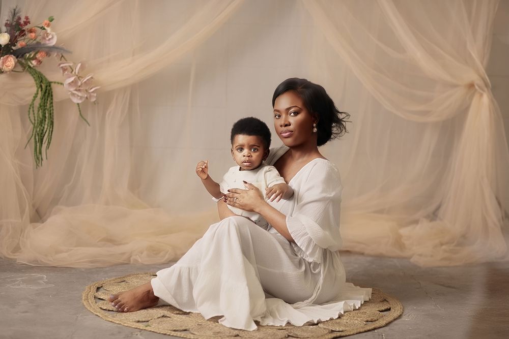 Mother in white dress sits on woven mat holding baby, posed before sheer cream draping in elegant studio portrait.