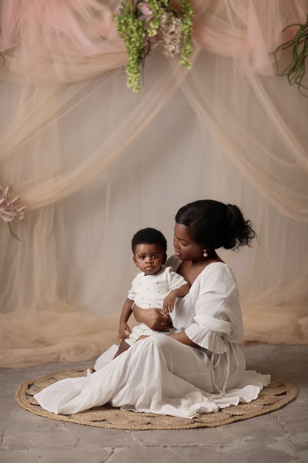 A Black mother in white dress sits on woven rug holding her baby, surrounded by soft tulle draping and floral decor.