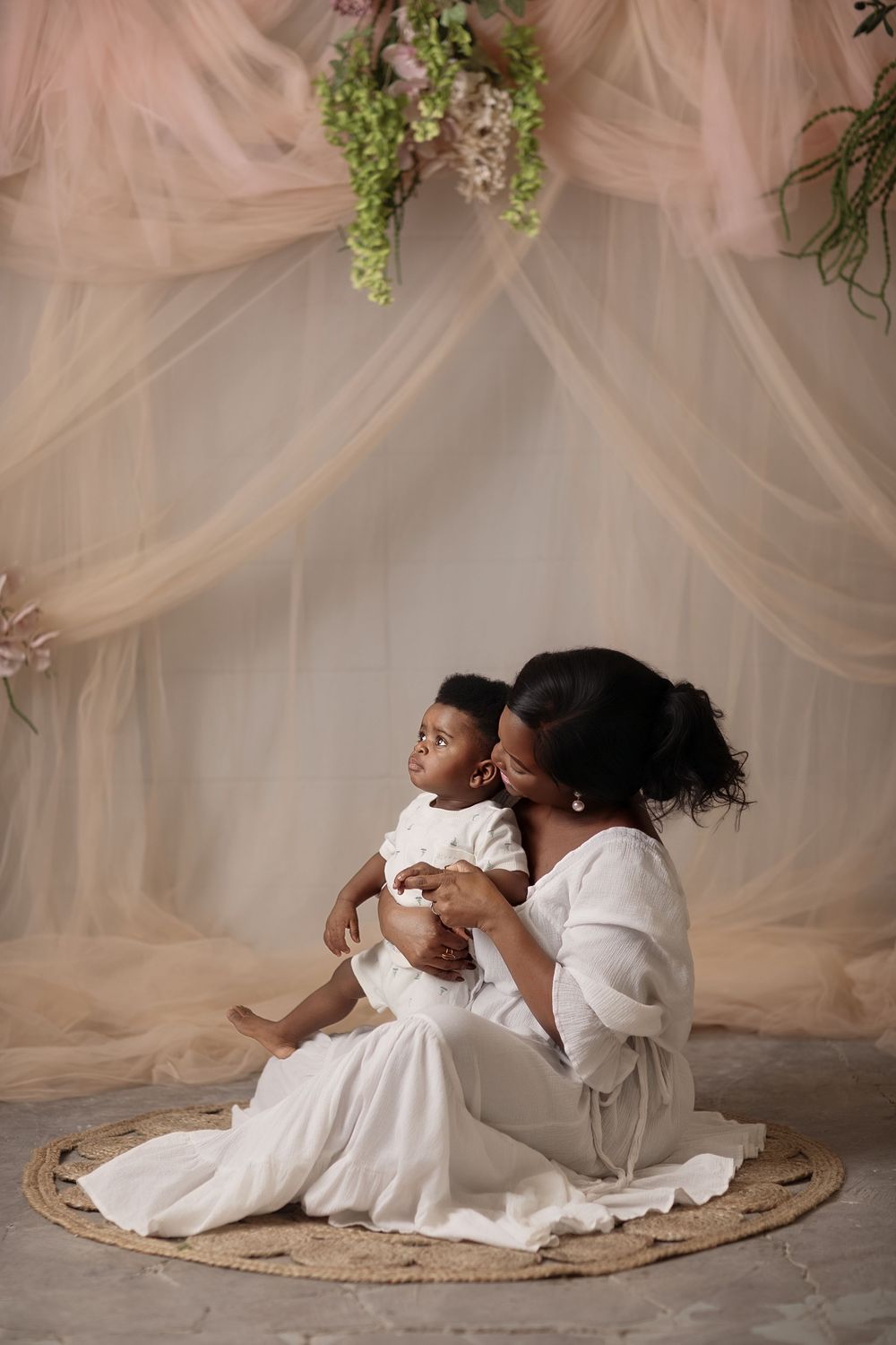 A mother in a white dress sits on a round rug holding her baby, surrounded by soft pink draping and floral decor.