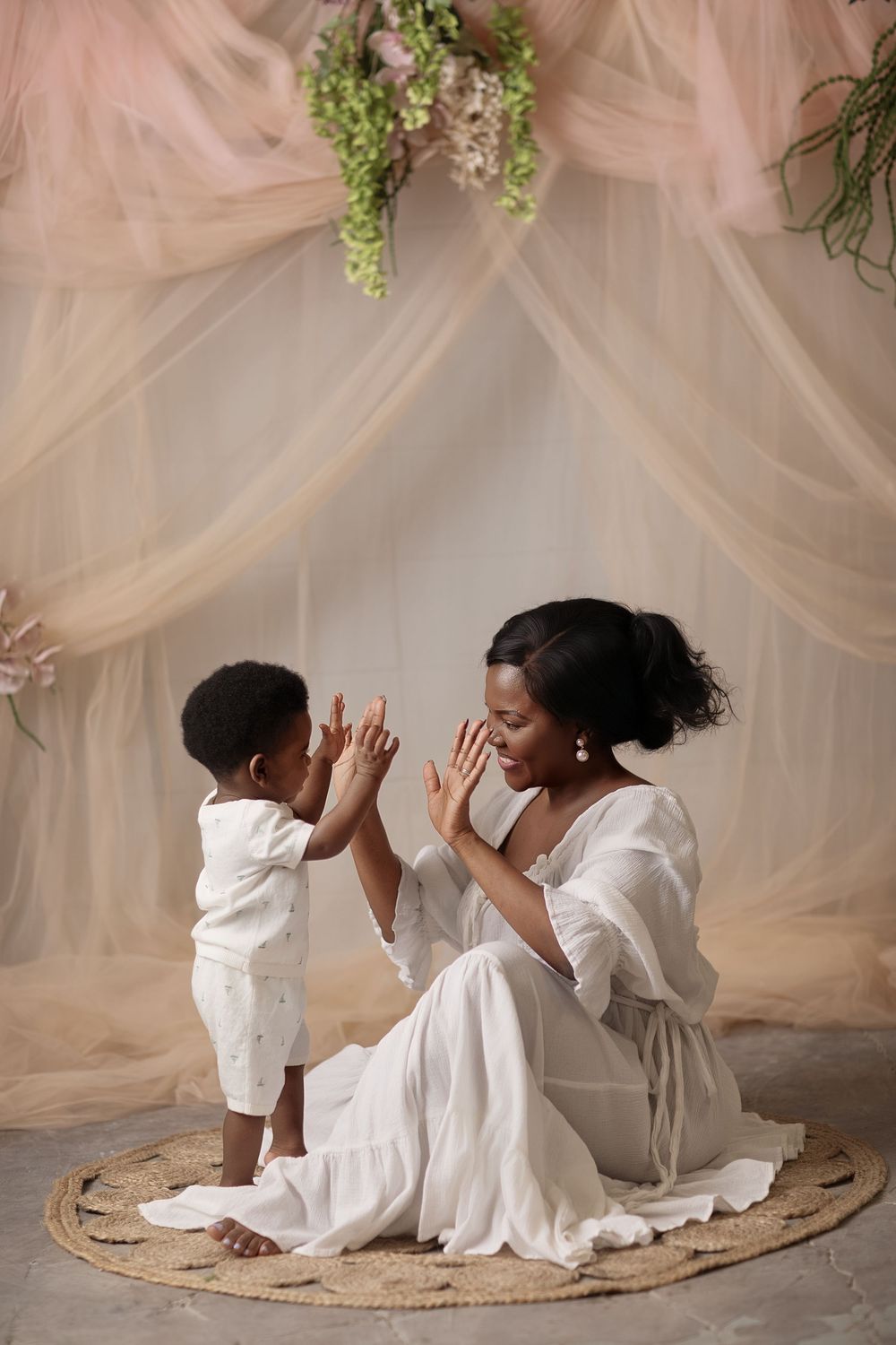 Black mother in white dress plays with toddler under floral canopy in elegant studio setting.
