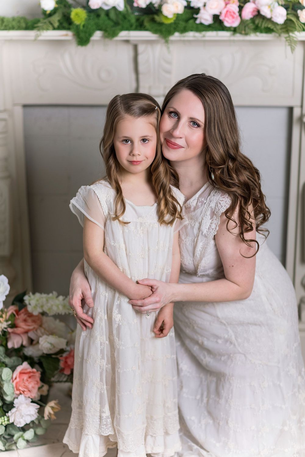 A mother and daughter in matching white dresses pose together near floral decorations in an elegant indoor setting.
