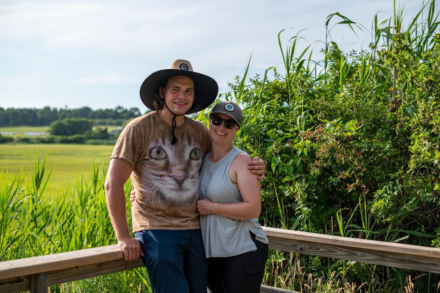 Couple smiling together on a wooden bridge overlooking a lush green marsh on a sunny day.