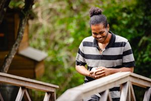 Smiling man with hair bun using smartphone on wooden bridge surrounded by lush green trees outdoors.