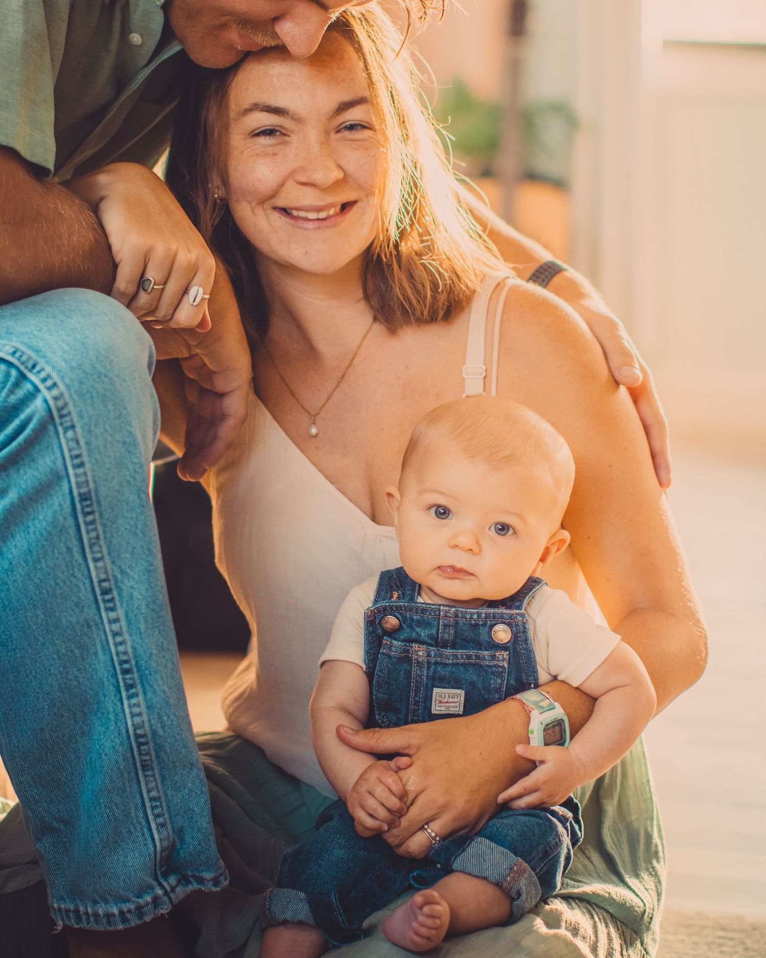 Smiling young woman holding a baby in denim overalls, with a man beside her in a warm, golden-lit family portrait.