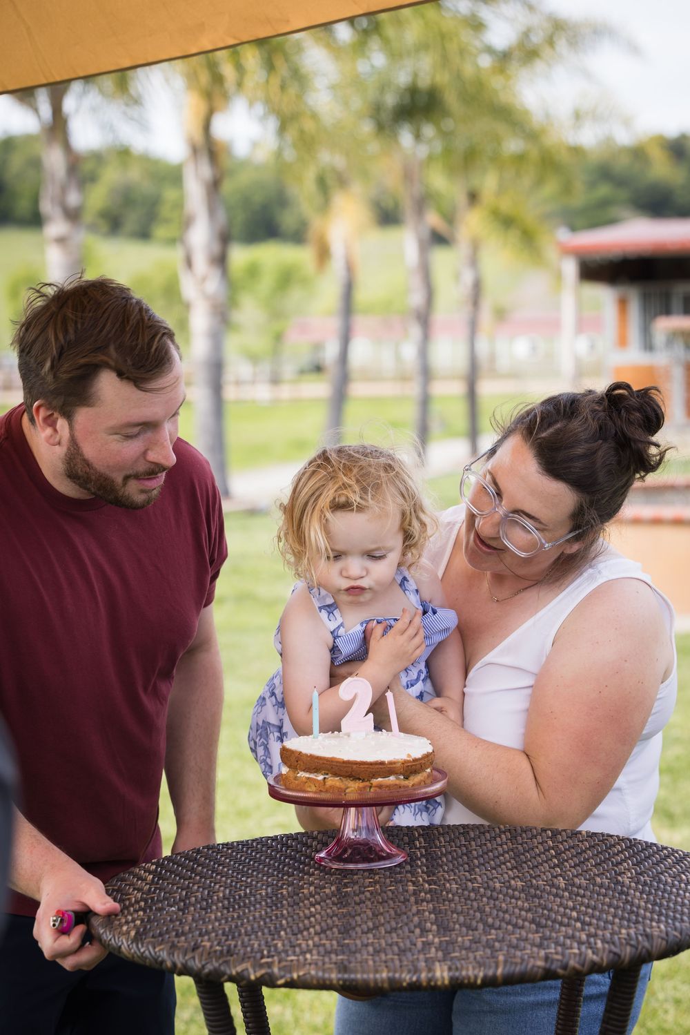 Family celebrating toddler's 2nd birthday with cake outdoors, parents smiling at child near wicker table.