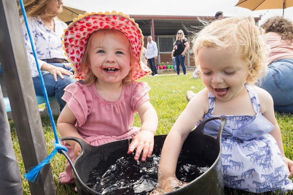 Two laughing toddler girls playing in a water tub at an outdoor event on a sunny day.