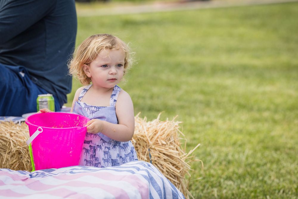Toddler girl in floral dress holding pink bucket on hay bale at outdoor picnic on green grass.